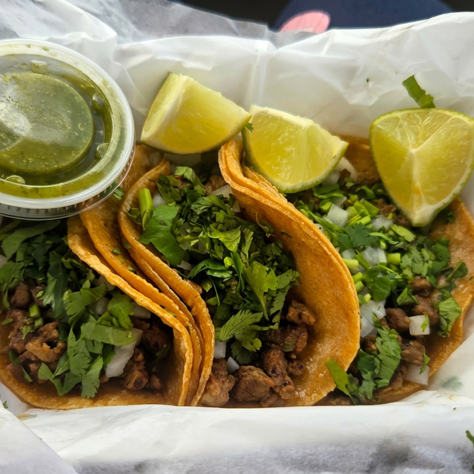 Three tacos filled with grilled meat, cilantro, onions, and lime wedges, served with green salsa in a paper-lined tray.